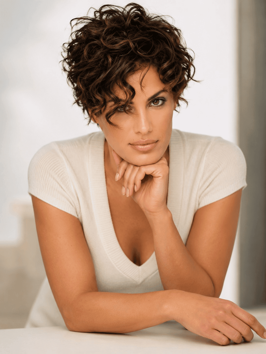 Woman with short curly hair and a thoughtful pose, wearing a white top near a bright window.