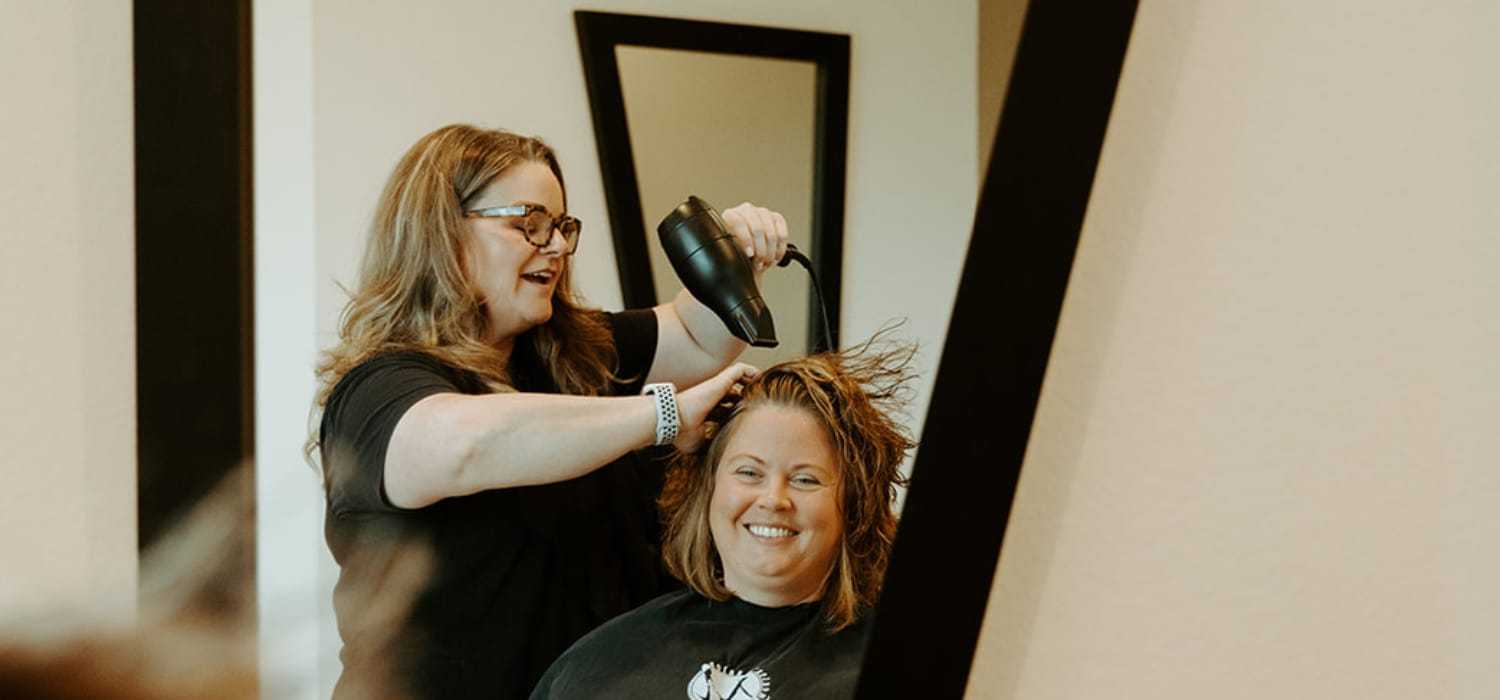 Hairdresser blow-drying a smiling woman's hair in a salon.