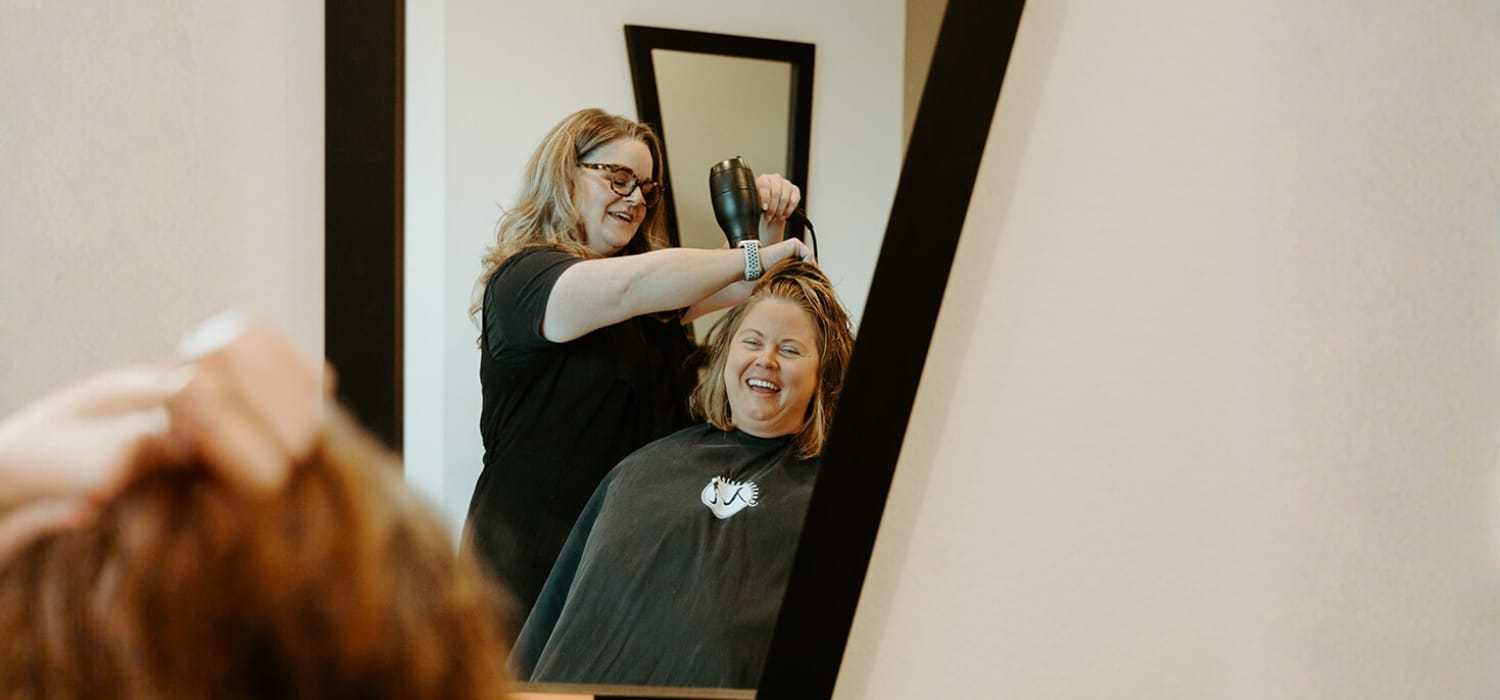Hairdresser blow-drying a client's hair in front of a mirror.